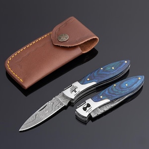 May include: Two folding knives with blue and gold handles and patterned blades, and a brown leather sheath. The knives have a tree logo and text on the blade. The sheath has a star-shaped button.