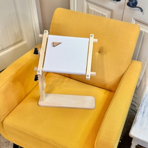 Peut inclure: Un support de cercle à broder en bois avec un tissu blanc tendu. Le support est en bois clair et possède des boutons réglables. Un petit fanion en bois est fixé au tissu. Le support est posé sur un fauteuil jaune.