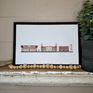 May include: A framed cross-stitch artwork depicting a cityscape with five buildings in a row. The buildings are white with red roofs and are set against a white background. The artwork is framed in black.