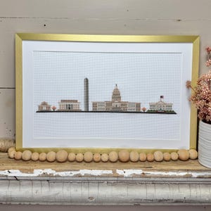 May include: Cross-stitch artwork of a city skyline, framed in gold. The artwork depicts several buildings, including a tall obelisk and a domed structure, likely representing Washington D.C. The piece is displayed on a distressed, white-painted wooden surface.