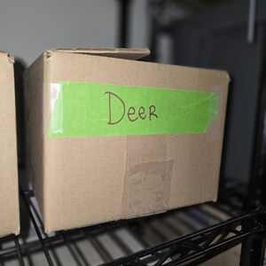 May include: A brown cardboard box with a green label that says "Deer" on it. The box is sitting on a black metal shelf.