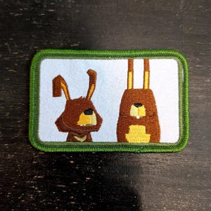 Op de afbeelding: Een rechthoekige patch met twee cartoon konijnen. De konijnen zijn bruin met gele details, tegen een lichtblauwe achtergrond. De patch heeft een groene rand. Waarschijnlijk een opstrijk- of opnaaipatch voor kleding of accessoires.