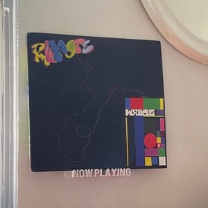 May include: A black record player with a pink vinyl record on the turntable. The record player has a silver tone arm and a black control panel. The record player is sitting on a black surface. A black and white album cover with the text "RIMOSTE" and "NOW PLAYING" is on the wall behind the record player.