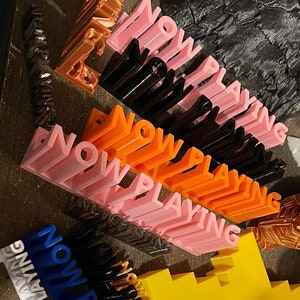 May include: A collection of 3D printed "NOW PLAYING" signs in various colors, including pink, orange, black, white, and blue. The signs are arranged in a chaotic manner on a surface with other 3D printed objects.