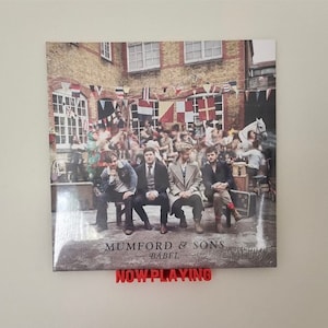 May include: A vinyl record album cover featuring a band of five men sitting on a bench in front of a brick building. The album cover is for Mumford & Sons' album "Babel". The album cover is displayed on a wall with a red "NOW PLAYING" sign below it.