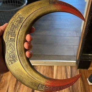 May include: A crescent-shaped prop with a gold and red color scheme. The prop has a curved design with intricate carvings along the edges. The tips are painted red, and the main body is gold. The prop is held in a hand.
