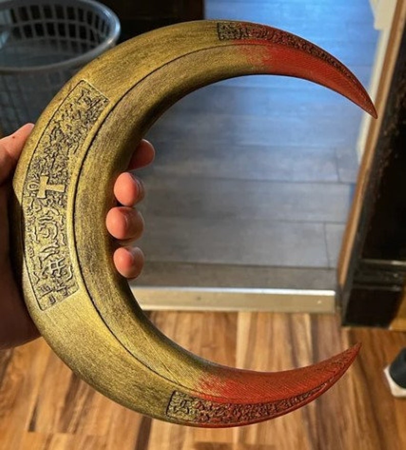 May include: A crescent-shaped prop with a gold and red color scheme. The prop has a curved design with intricate carvings along the edges. The tips are painted red, and the main body is gold. The prop is held in a hand.