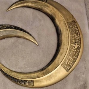 May include: Two curved, crescent-shaped blades with a metallic gold finish. The blades have a sharp point and are decorated with intricate, dark etchings. The blades are likely props or decorative items.