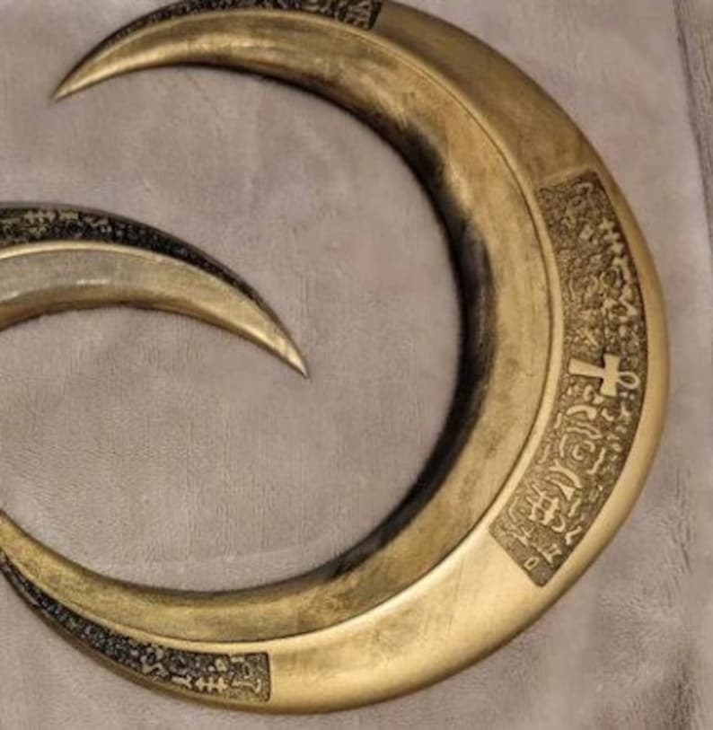 May include: Two curved, crescent-shaped blades with a metallic gold finish. The blades have a sharp point and are decorated with intricate, dark etchings. The blades are likely props or decorative items.