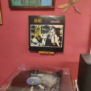 May include: A vinyl record by Tom Waits titled "Swordfishtrombones" with the text "Now Playing" displayed below the album cover. The album cover features a black and white image of three men in a dimly lit room.