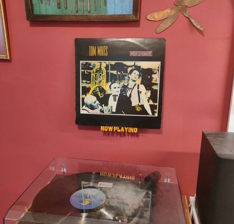 May include: A vinyl record by Tom Waits titled "Swordfishtrombones" with the text "Now Playing" displayed below the album cover. The album cover features a black and white image of three men in a dimly lit room.