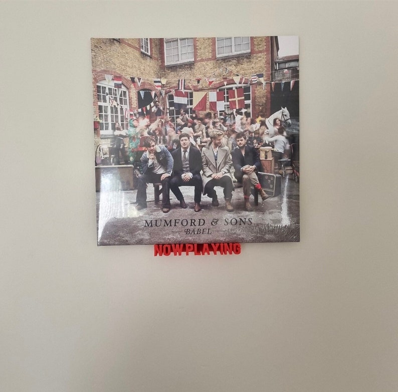 May include: A vinyl record album cover featuring a band of five men sitting on a bench in front of a brick building. The album cover is for Mumford & Sons' album "Babel". The album cover is displayed on a wall with a red "NOW PLAYING" sign below it.