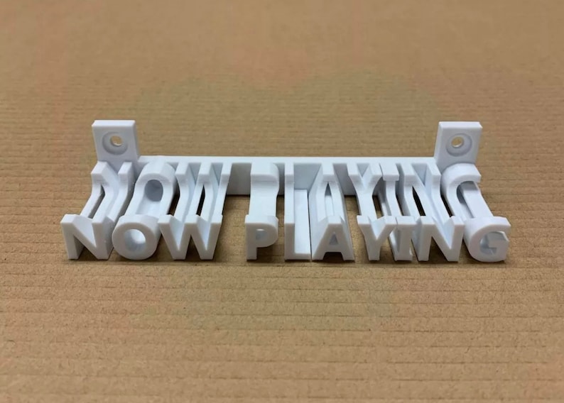 May include: White plastic sign with the words "NOW PLAYING" in 3D letters. The sign is mounted on a wall with two small brackets.