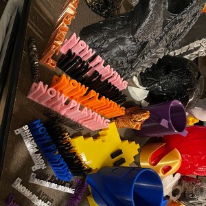 May include: A collection of 3D printed objects, including a Pac-Man character, a blue and white cone, a purple cup, and several orange, pink, and black signs that read "NOW PLAYING".