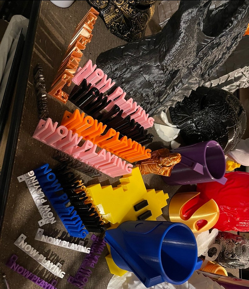 May include: A collection of 3D printed objects, including a Pac-Man character, a blue and white cone, a purple cup, and several orange, pink, and black signs that read "NOW PLAYING".