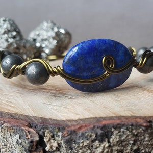 May include: A wire-wrapped bracelet with a large oval lapis lazuli stone and smaller round hematite beads. The bracelet is made with brass wire and has a simple, elegant design.