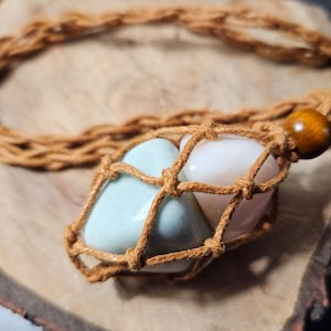 May include: A brown macrame necklace with a wooden bead and three heart-shaped stones in a net. The stones are light blue, pink, and white.