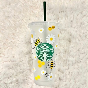 May include: A clear plastic reusable cup with a white and green Starbucks logo, a bee and honeycomb design, and a green straw. The cup is decorated with white daisies.