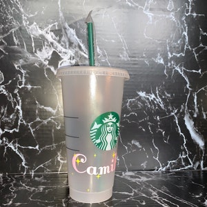 May include: A clear plastic reusable cup with a green Starbucks logo and the name "Camil" written in pink with yellow stars. The cup has a green straw.