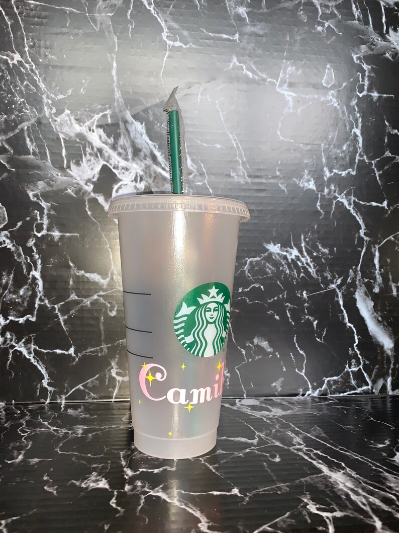 May include: A clear plastic reusable cup with a green Starbucks logo and the name "Camil" written in pink with yellow stars. The cup has a green straw.