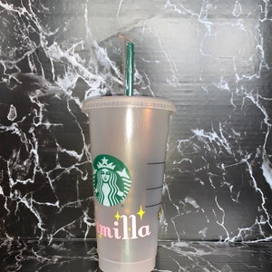 May include: A clear plastic reusable cup with a green Starbucks logo and the name "Camilla" written in pink letters with yellow stars. The cup has a green straw.