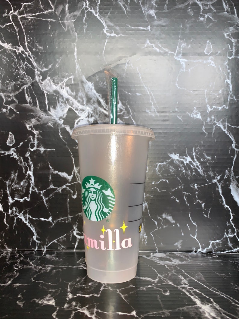 May include: A clear plastic reusable cup with a green Starbucks logo and the name "Camilla" written in pink letters with yellow stars. The cup has a green straw.