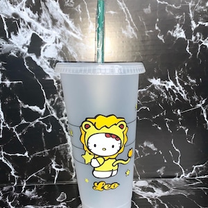 May include: A clear plastic reusable cup with a black lid and a green straw. The cup has a Hello Kitty design with Hello Kitty dressed as a lion. The design includes the words "Leo" and stars.