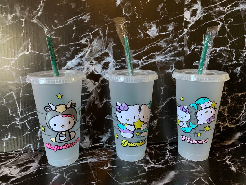 May include: Three clear plastic cups with green straws and Hello Kitty designs. Each cup features a different zodiac sign: Capricorn, Gemini, and Pisces.