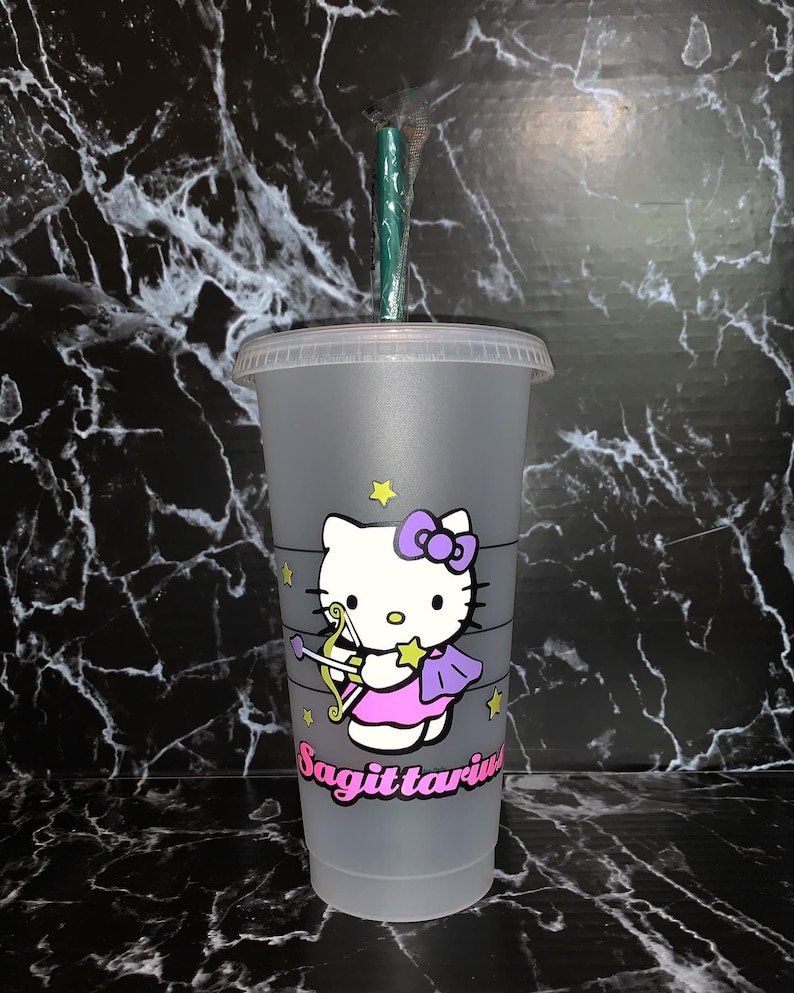 May include: A gray reusable cup with a Hello Kitty design featuring Sagittarius. The cup has a green straw and a white lid. The Hello Kitty character is wearing a pink dress and purple wings with a bow on her head. The word "Sagittarius" is written in black below the character.