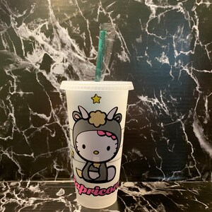 May include: White reusable cup with a Hello Kitty design. The design features Hello Kitty dressed as a Capricorn with a pink star on the cup.