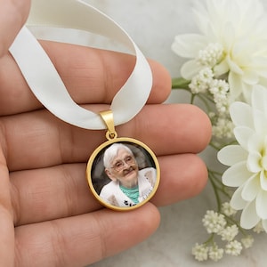 May include: A gold-colored pendant with a photo of an elderly person, attached to a white ribbon. The pendant is round and has a gold-colored border. The ribbon is held by a hand, with white flowers in the background.