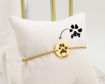 May include: Gold-colored bracelet with a circular charm featuring a paw print and the name "Tucker". The bracelet is displayed on a white pillow, with a paw print illustration above it.