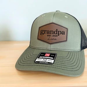 Puede incluir: Gorra de camionero verde y negra con un parche de cuero marrón que dice "grandpa EST. 2025 Luna". La gorra tiene una visera curva y una correa ajustable. Una etiqueta Richardson 112 Trucker es visible.