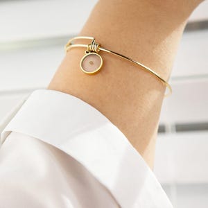 May include: Gold-coloured bangle bracelet featuring a circular charm. The charm has a pale pink backdrop and a tiny, tan seed at its centre. The bracelet is worn on an arm, with a white shirt visible.