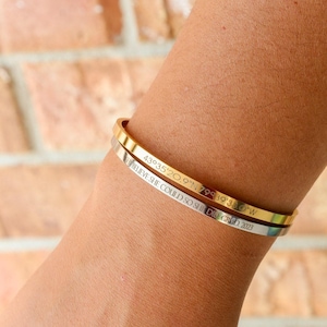 Puede incluir: Dos pulseras finas grabadas. Una es dorada, la otra plateada. La pulsera dorada tiene grabadas las coordenadas "43°35'20.9"N 79°39'35.0"W". La pulsera plateada dice "Believe she could so she did. Grad 2023."