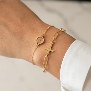 Könnte beinhalten: Zwei goldene Armbänder. Eines hat einen kleinen, runden Anhänger mit einem Samen darin. Das andere hat einen Kreuzanhänger und das eingravierte Datum "08-24-2024". Die Armbänder werden an einem Handgelenk getragen, mit einem weißen Hemd im Hintergrund.