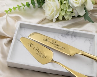May include: A gold-colored cake knife and server set with the inscription "The Bricenos June 2025" on each piece. The set rests on a white marble tray, with a bouquet of white roses in the background. Wedding and event decor.