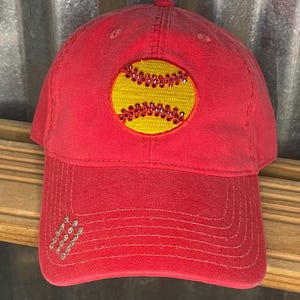 May include: Red baseball cap with a yellow softball design embellished with rhinestones. The cap has a curved brim and a series of rhinestones on the side. The cap is made of a soft, worn material.