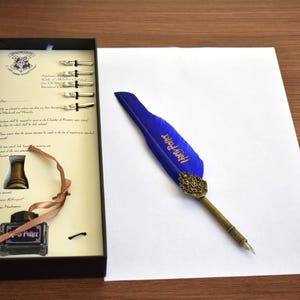 Könnte beinhalten: Eine blaue und goldene Federkiel mit den Worten "Harry Potter" darauf, eine kleine Tintenflasche und eine Reihe von Schreibfedern in einer schwarzen Schachtel. Die Schachtel trägt ein Hogwarts-Wappen und die Worte "Hogwarts Schule für Hexerei und Zauberei" sind darauf gedruckt.