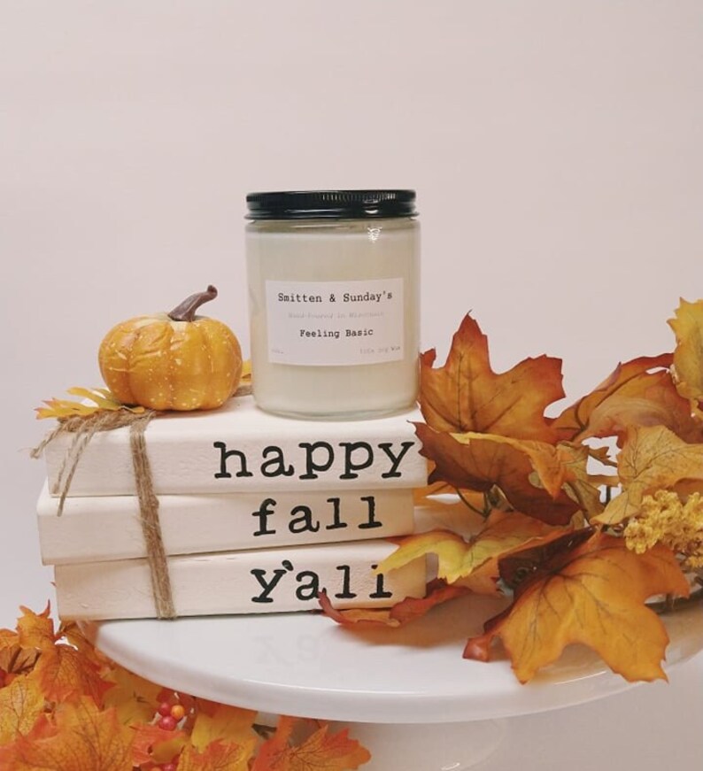 May include: A white candle jar with a black lid and the text "Smitten & Sunday's" and "Feeling Basic" on the label. The candle is sitting on a stack of white books with the text "happy fall y'all" on the front. The books are surrounded by fall leaves.