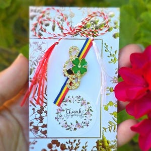 May include: A handmade greeting card with a gold-colored treble clef and green shamrock charm. The card features a floral design, a ribbon with red, yellow, and blue stripes, and the words "Thank you".