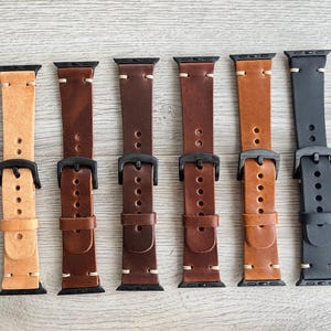 May include: A collection of six leather watch bands in various shades of brown and one black band. Each band features a black buckle and stitching, showcasing a range of colors from light tan to dark brown and black.
