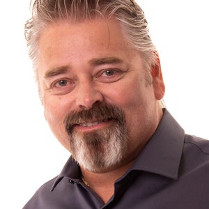 May include: A man with grey hair and a goatee beard is wearing a dark grey collared shirt. He is smiling and looking at the camera.