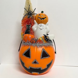 May include: Orange pumpkin shaped candy bucket with a black and silver glitter face. The bucket is filled with a white ghost, a black cat, a small orange pumpkin, a spider, and a candy corn tree.
