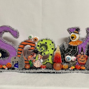 May include: A Halloween-themed decoration spelling out the word "SPOOKY" in purple, orange, and green letters. The letters are decorated with Halloween figures, pumpkins, and eyeballs. The base is trimmed with silver tinsel.