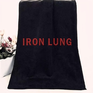 Puede incluir: Toalla negra con el texto bordado "IRON LUNG" en rojo. La toalla está doblada y parece estar hecha de un material suave y absorbente. El fondo es de color claro y a la izquierda se ve un pequeño jarrón con flores.