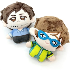 Puede incluir: Dos muñecos de peluche con cabello castaño y rasgos faciales bordados. Uno lleva una camisa azul y pantalones grises, el otro un mono verde y amarillo y gafas azules. Ambos muñecos tienen orejas pequeñas sobre un fondo blanco.