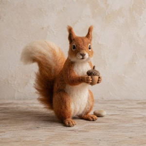 Puede incluir: Una figura de ardilla de lana fieltrada con pelaje marrón rojizo, pecho color crema y una gran cola tupida. La ardilla está de pie y sostiene una pequeña bellota marrón en sus patas delanteras, con una pequeña piedra lisa al lado.