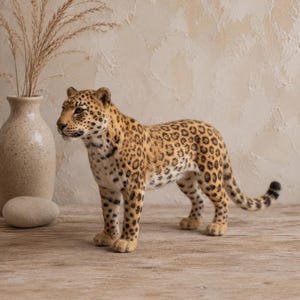 Könnte beinhalten: Eine nadelgefilzte Leopardenfigur mit einem goldbraunen Fell, das mit dunkelbraunen Rosetten und Flecken gemustert ist. Der Leopard steht in einer Seitenansicht, wobei der Kopf leicht zum Betrachter gedreht ist, und zeigt einen langen Schwanz.