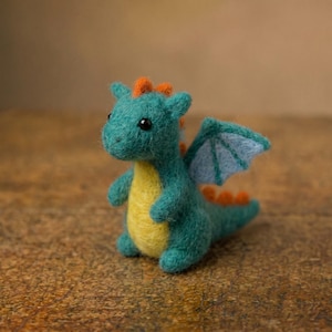May include: A needle-felted dragon figurine featuring a teal body, a bright yellow belly, and orange spikes along its back. The creature has small, light blue wings and black bead eyes, standing in an upright position with a rounded, soft texture.